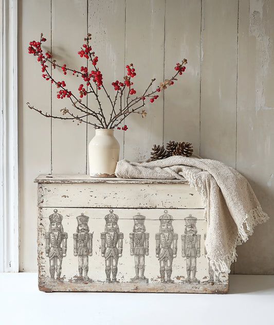Decorative chest with nutcracker design using Iron Orchid Design Stamps, white vase with red branches, and beige blanket against a wooden wall.