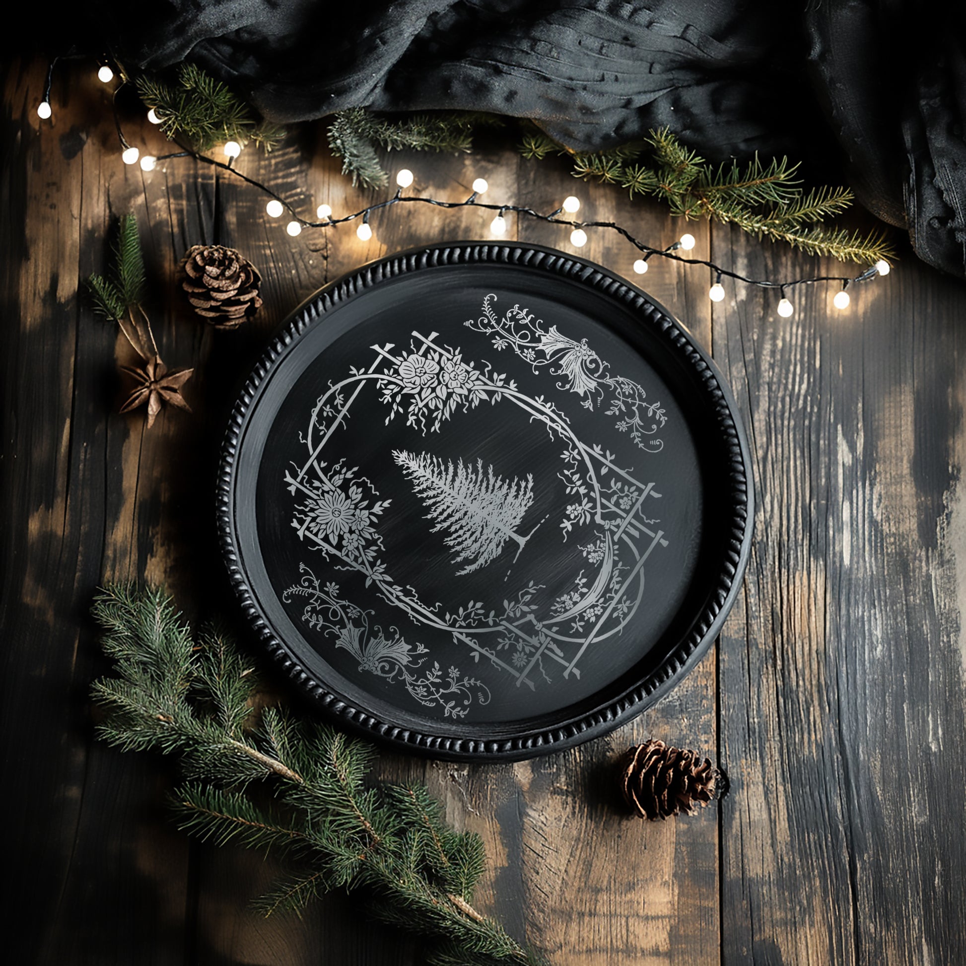 Decorative black plate with white floral design on a wooden surface with pine branches and lights.