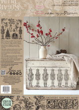 Load image into Gallery viewer, The back cover of IOD Stamp Sugar Plum featuring decorative chest with Nutcracker design, white vase with red flowers, and wooden wall background.