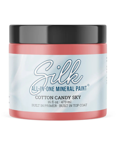 A jar of Silk All-In-One Mineral Paint in the color Cotton Candy Sky, featuring a light blue and white label with blue text, a coral pink base, and a black lid.