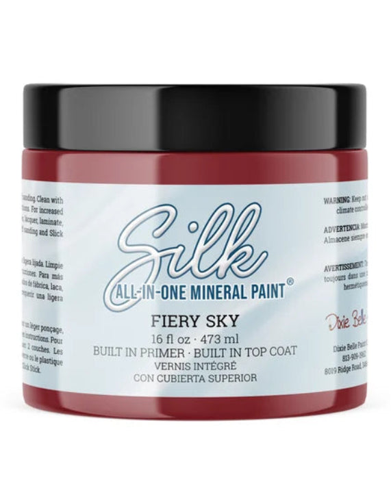 A jar of Silk All-In-One Mineral Paint in the shade Fiery Sky, featuring a deep red paint visible through the clear container, a black lid, and a white label with blue and red text.