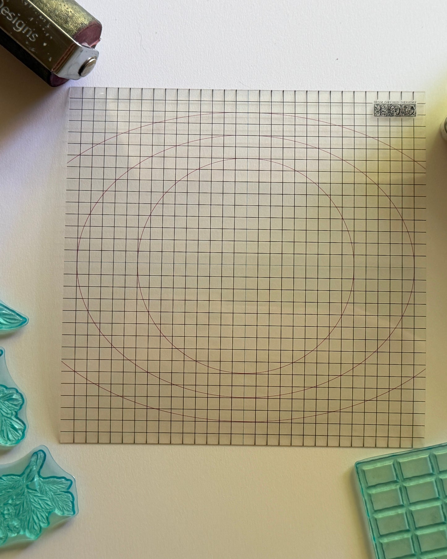 A clear acrylic gridded square template with circular guides, shown on a white surface alongside craft tools and rubber stamps.