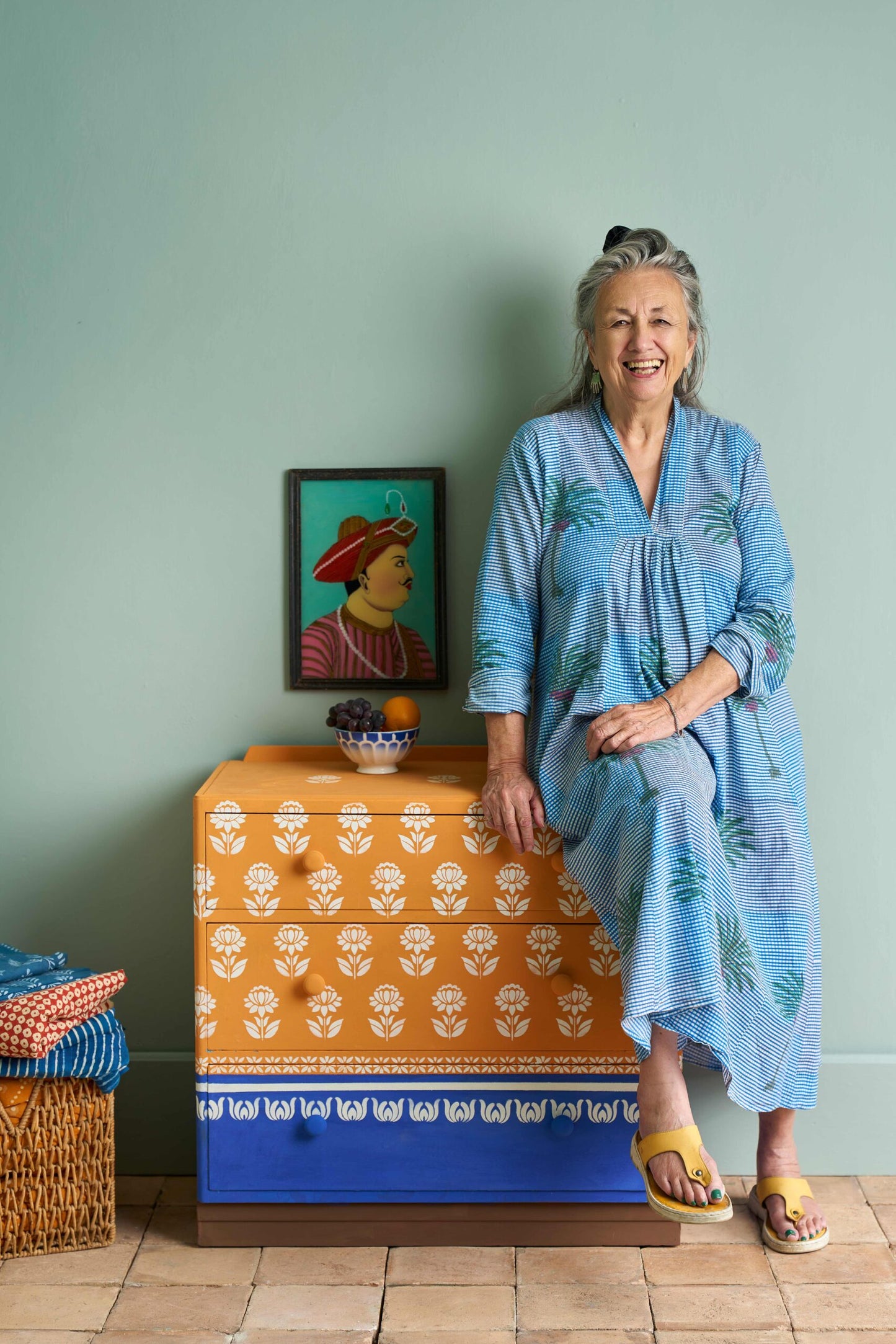 Annie Sloan in a blue dress standing next to a colourful dresser with a painting on the wall.