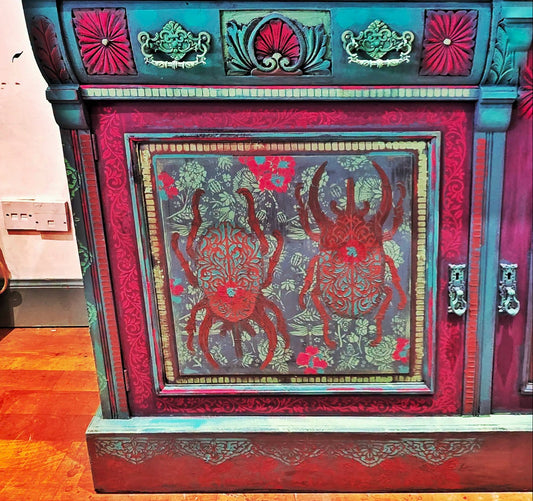 A highly decorative, hand-painted wooden cabinet featuring a central panel with two red bugson a blue-gray background. The cabinet has a vibrant teal and red painted frame with intricate floral patterns and ornate metal handles, set against a home interior background