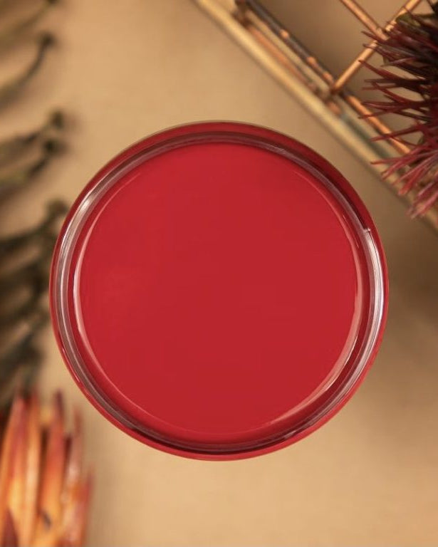 A round glass jar filled with vibrant red paint, shown from a top-down view on a beige background with decorative dried botanicals.